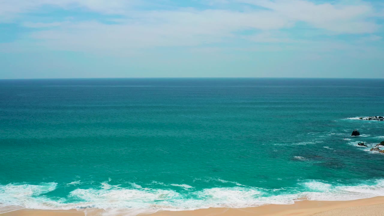 At 0:02, blue-green water dominates the Secrets Marquis Los Cabos frame, with foamy surf spread across the lower edge, small rock outcrops to the right, and no visible people.