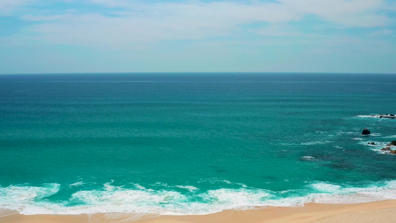 Mid-clip, the elevated view over Secrets Marquis Los Cabos keeps the horizon level, with calm turquoise water, a white shore break across the lower frame, rocks at right, and no visible people.