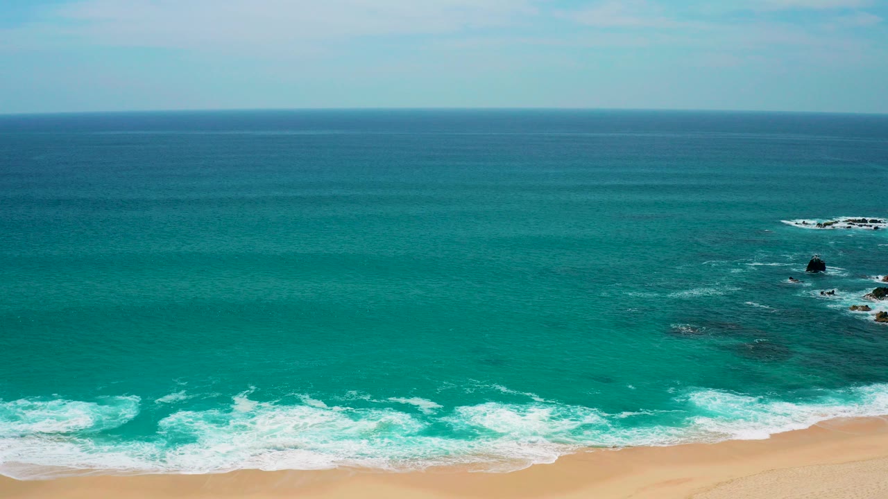Elevated view of turquoise ocean meeting tan sand at Secrets Marquis Los Cabos, Mexico, with dark rocks offshore and no visible people.