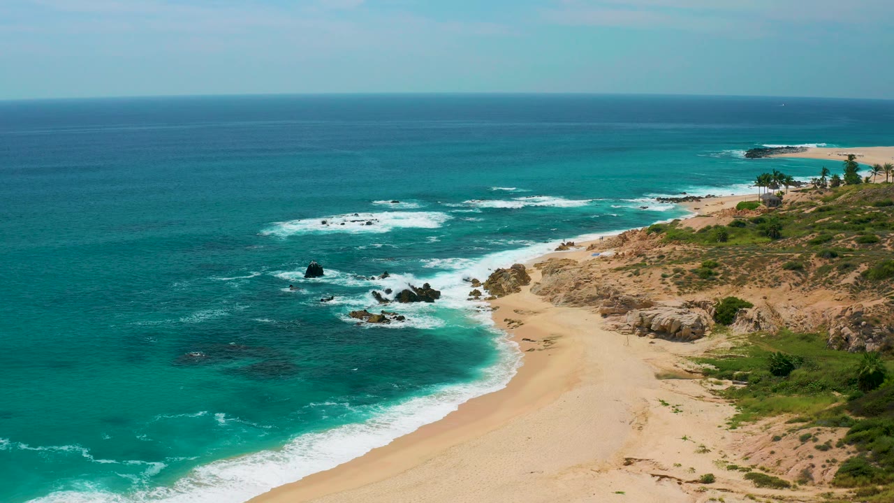 High coastal view at Secrets Marquis Los Cabos, Mexico with the ocean spread across the frame, surf on the beach, rocky terrain to the right, palms in the distance, and no visible people.