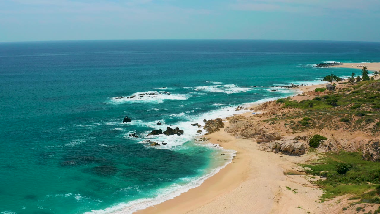 Aerial frame over Secrets Marquis Los Cabos, Mexico showing turquoise water, white surf, golden sand, dark rocks, and no visible people at the start of the clip.