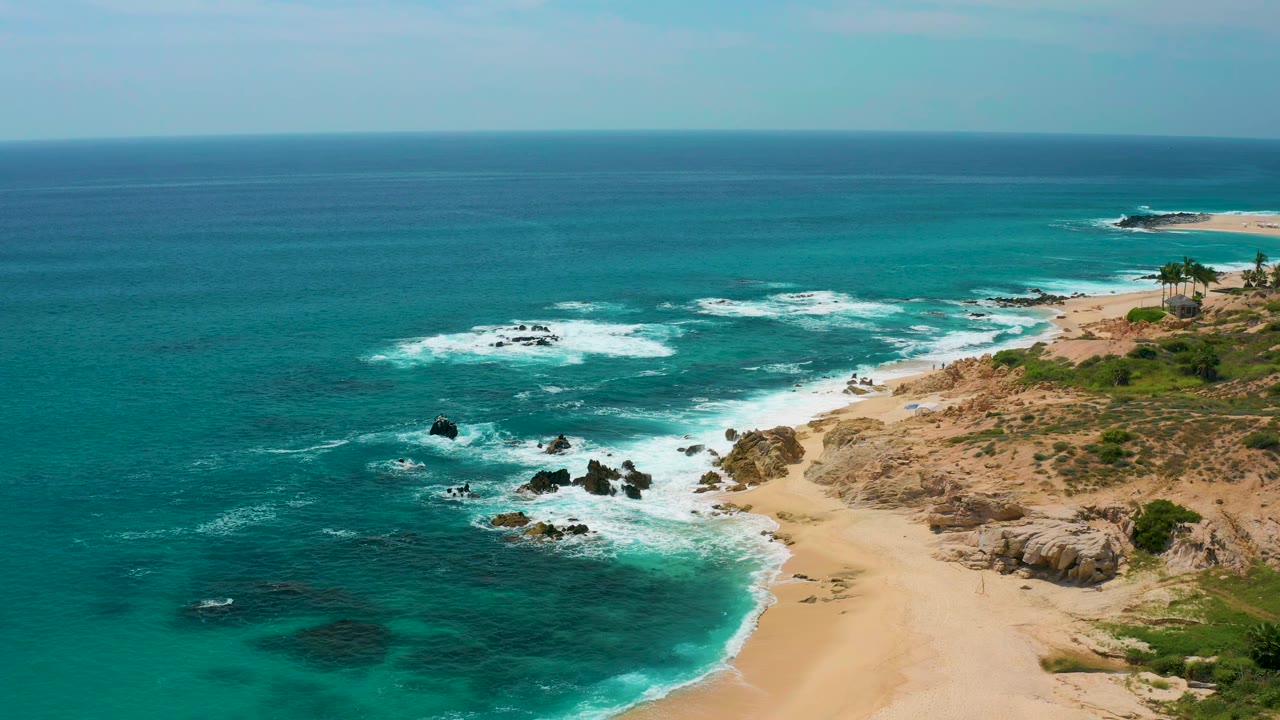 Aerial coastal frame at Secrets Marquis Los Cabos, Mexico, with white foam gathering around offshore rocks and a sandy beach curving along the right side, no visible people.