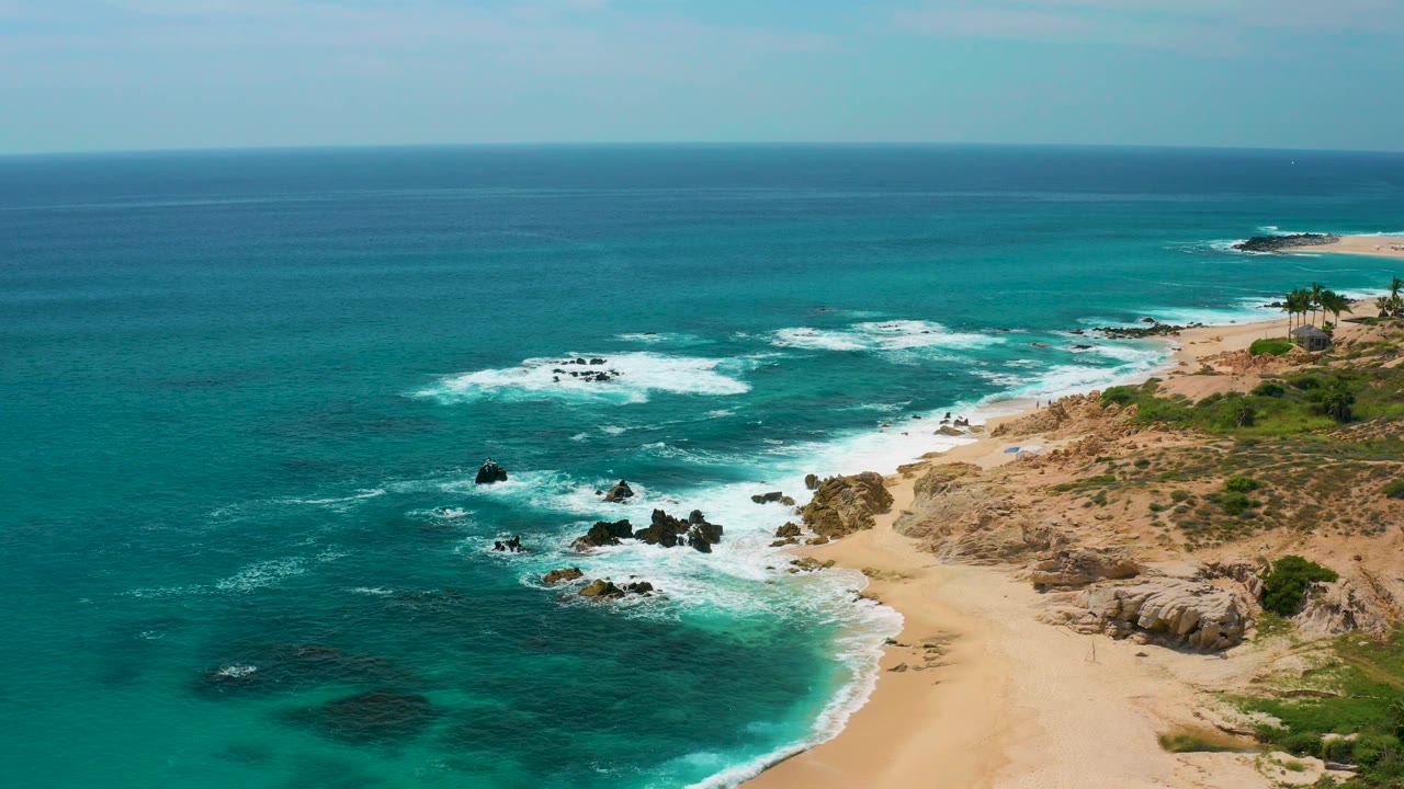 High ocean view of the Secrets Marquis Los Cabos, Mexico shoreline, where surf lines move toward tan sand, dark rocks, and scrubby coastal plants, with no visible people.