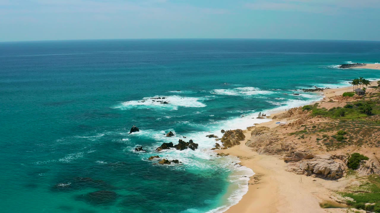 Wide elevated view at Secrets Marquis Los Cabos, Mexico, showing turquoise water, broken white surf, dark rock points, and an empty sandy shore with no visible people.