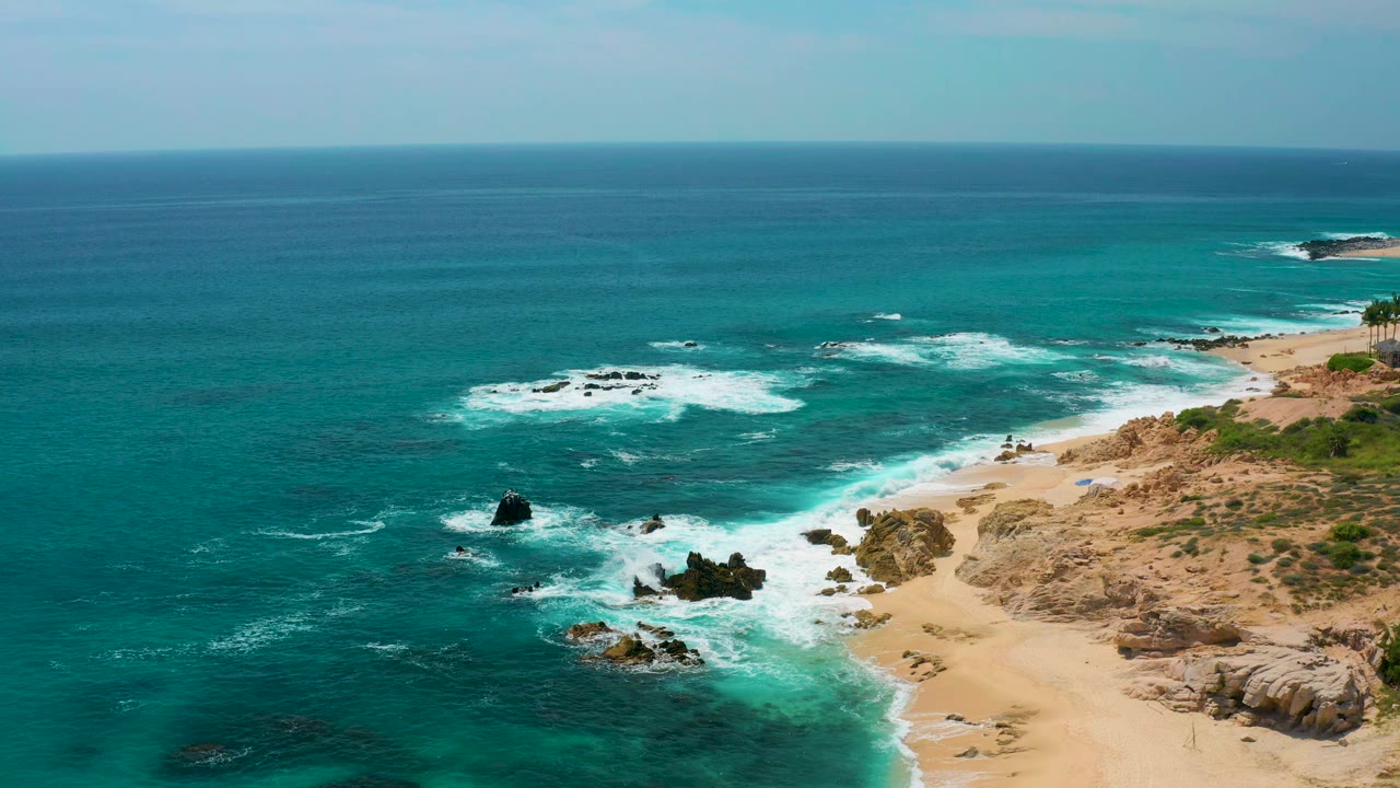 Elevated view at Secrets Marquis Los Cabos, Mexico, where white foam wraps around black coastal rocks beside a pale sandy beach, with no visible people.