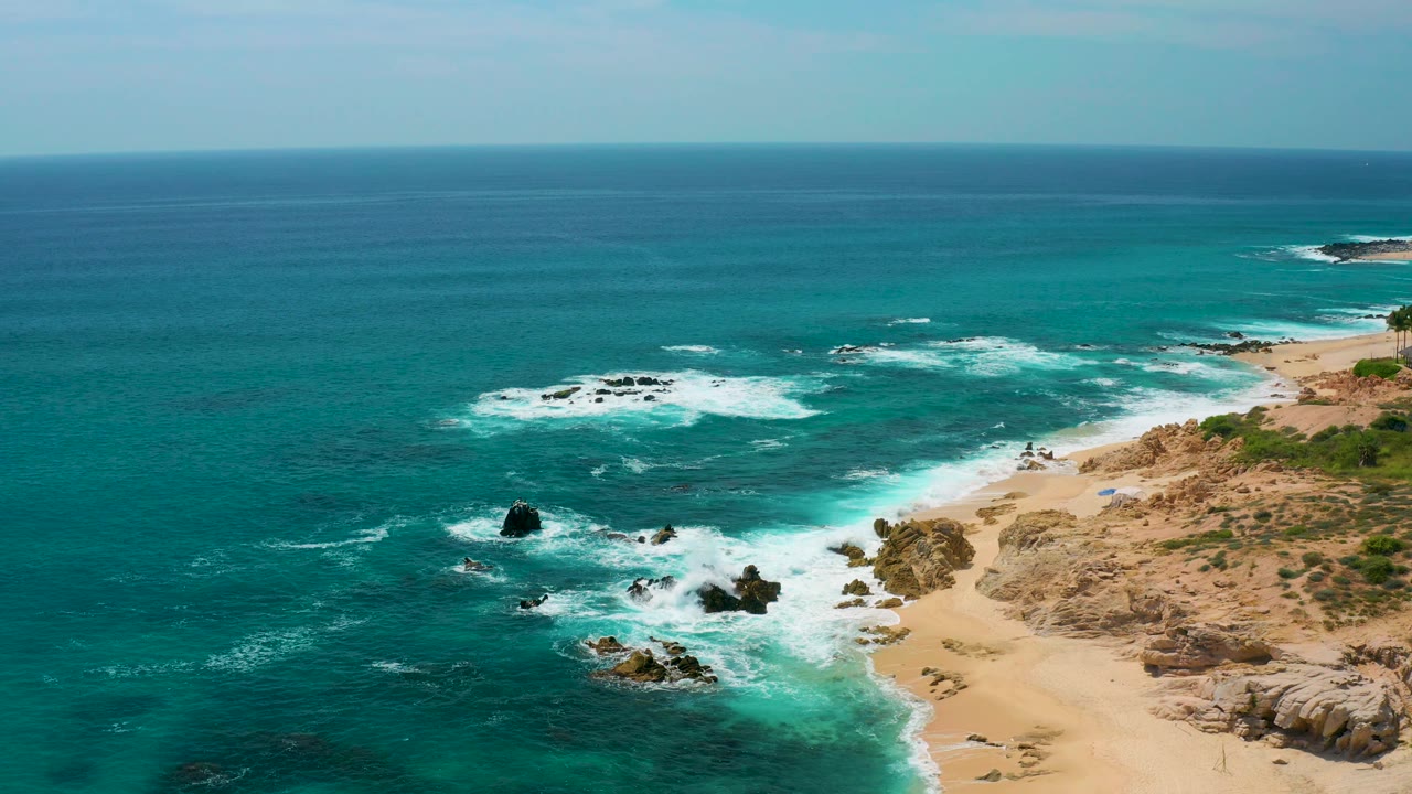 Wide aerial frame at Secrets Marquis Los Cabos, Mexico, with broad teal water, scattered foam lines, rocky sand shoreline to the right, and no visible people.