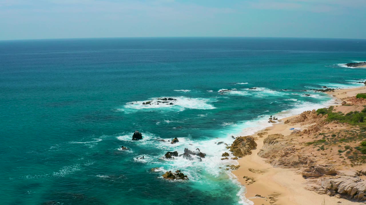 Drone-style view at Secrets Marquis Los Cabos, Mexico, showing surf breaking over several dark rocks with tan beach and rough shoreline terrain on the right, no visible people.