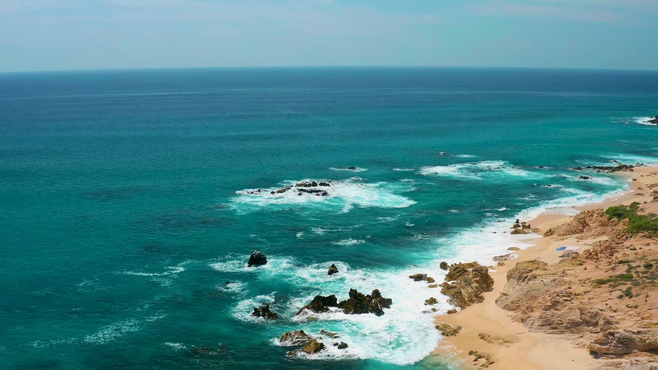 A wide aerial view at Secrets Marquis Los Cabos shows turquoise ocean, white surf around dark rocks, tan beach at right, and no visible people.