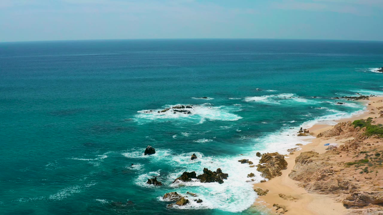 At 0:01, whitewater spreads wider around the central rocks while the sandy shoreline at Secrets Marquis Los Cabos remains along the right edge with no visible people.