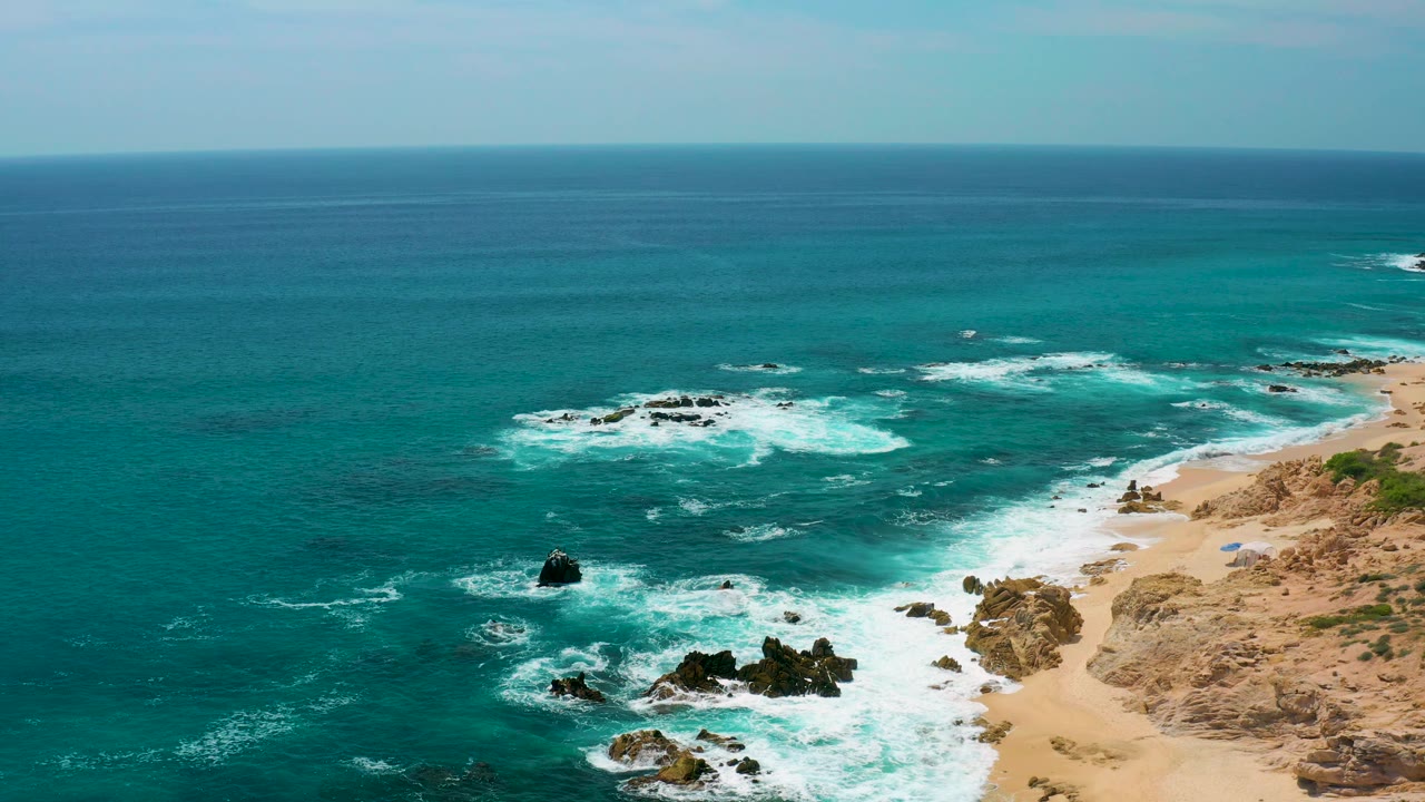At 0:02, the surf forms bright bands around several dark rock clusters, with open blue-green water offshore and no visible people on the beach or rocks.