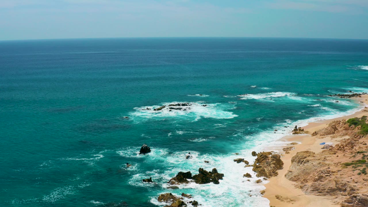 At 0:03, the camera view holds a broad horizon as white waves wash toward the sandy beach and dark rocks at Secrets Marquis Los Cabos, with no visible people.