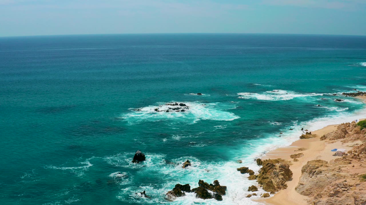 Aerial view at Secrets Marquis Los Cabos, Mexico, with turquoise water, white surf around offshore rocks, sandy shore on the right, and no visible people.