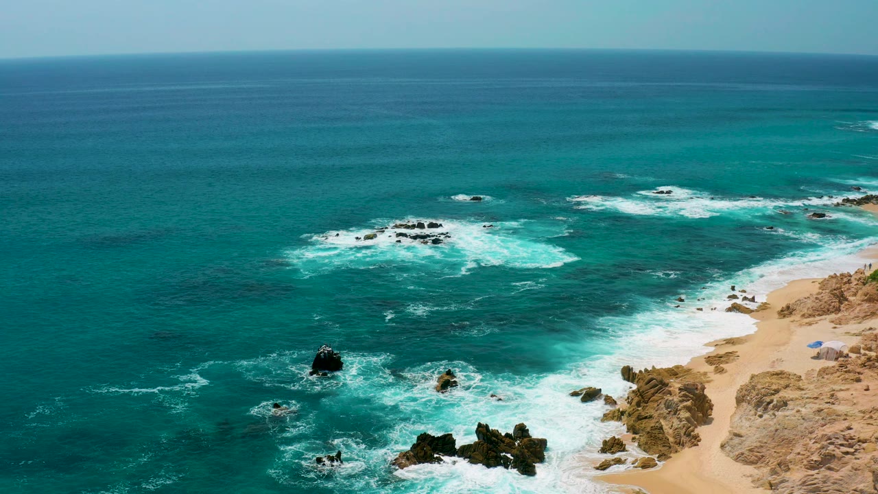 Wide aerial frame at Secrets Marquis Los Cabos, Mexico, with clear teal ocean, foam lines around rocks, sandy shoreline on the right, and no visible people.