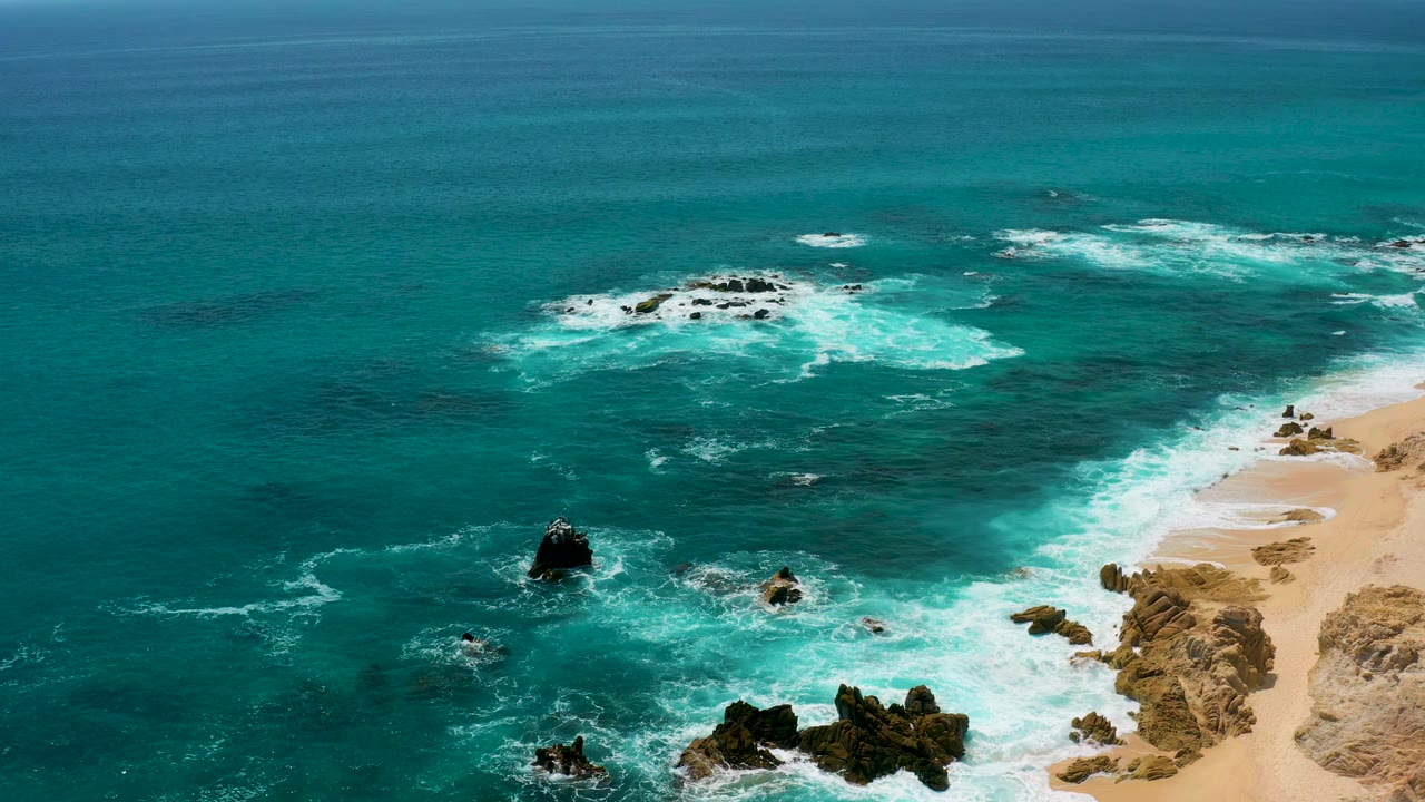 At 0:02, the drone perspective keeps open blue-green water across the top, foamy surf over scattered rocks, beige coastal rocks at right, and no visible people.