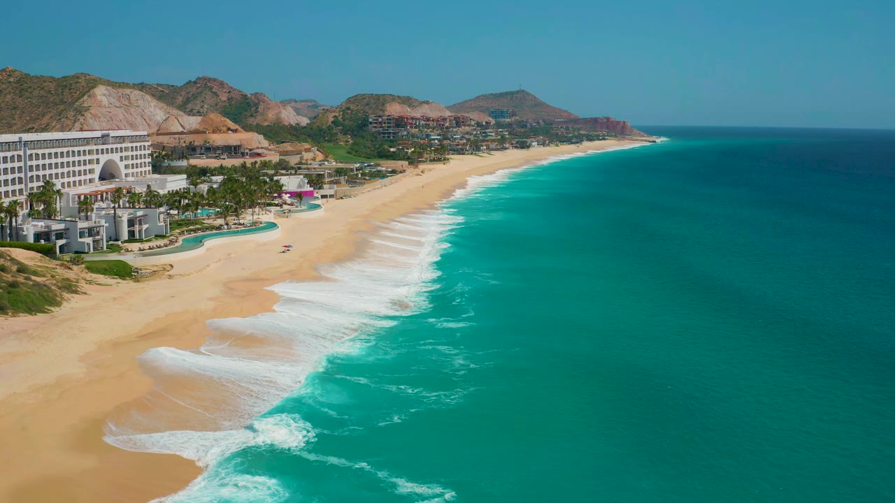 Aerial frame at Secrets Marquis Los Cabos, Mexico, showing the resort edge on the left, fresh whitewater spreading across the sand, open turquoise ocean to the right, and no visible people.
