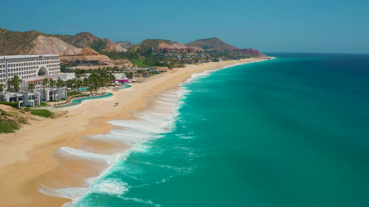 A wide aerial view of Secrets Marquis Los Cabos in Mexico, with the resort cropped along the left edge, stronger white surf bands on the sand, open ocean filling the right side, and no visible people.