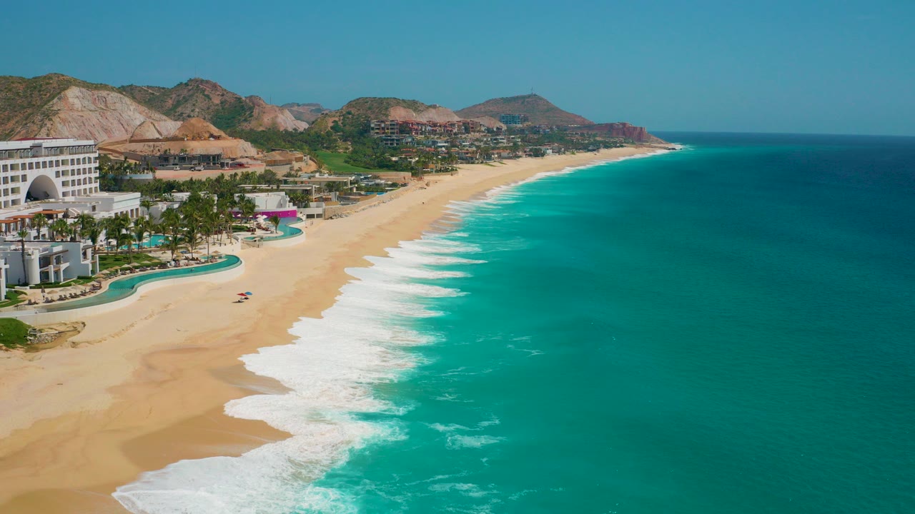 Start of the clip over Secrets Marquis Los Cabos in Mexico, with turquoise water, white surf, pale sand, resort pools at left, dry hills in back, and no visible people.