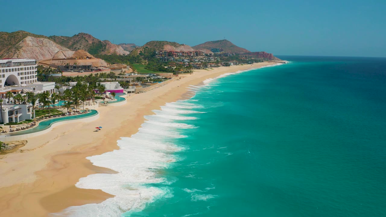 At 0:02, the aerial view holds the Mexico resort coastline with more open water at right, resort structures at left, foamy shorebreak, distant hills, and no visible people.