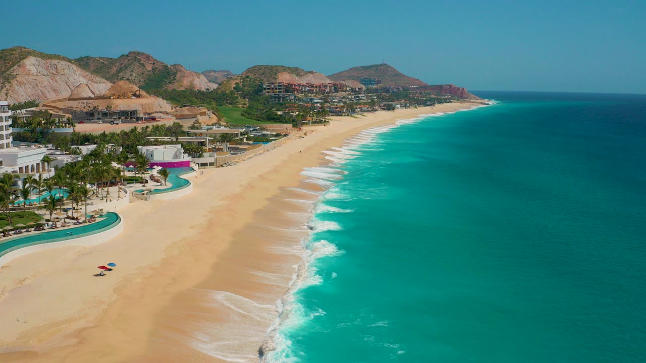 Aerial view at Secrets Marquis Los Cabos, Mexico, showing turquoise surf along a wide sandy beach with resort pools at left, mountains behind, and no visible people.
