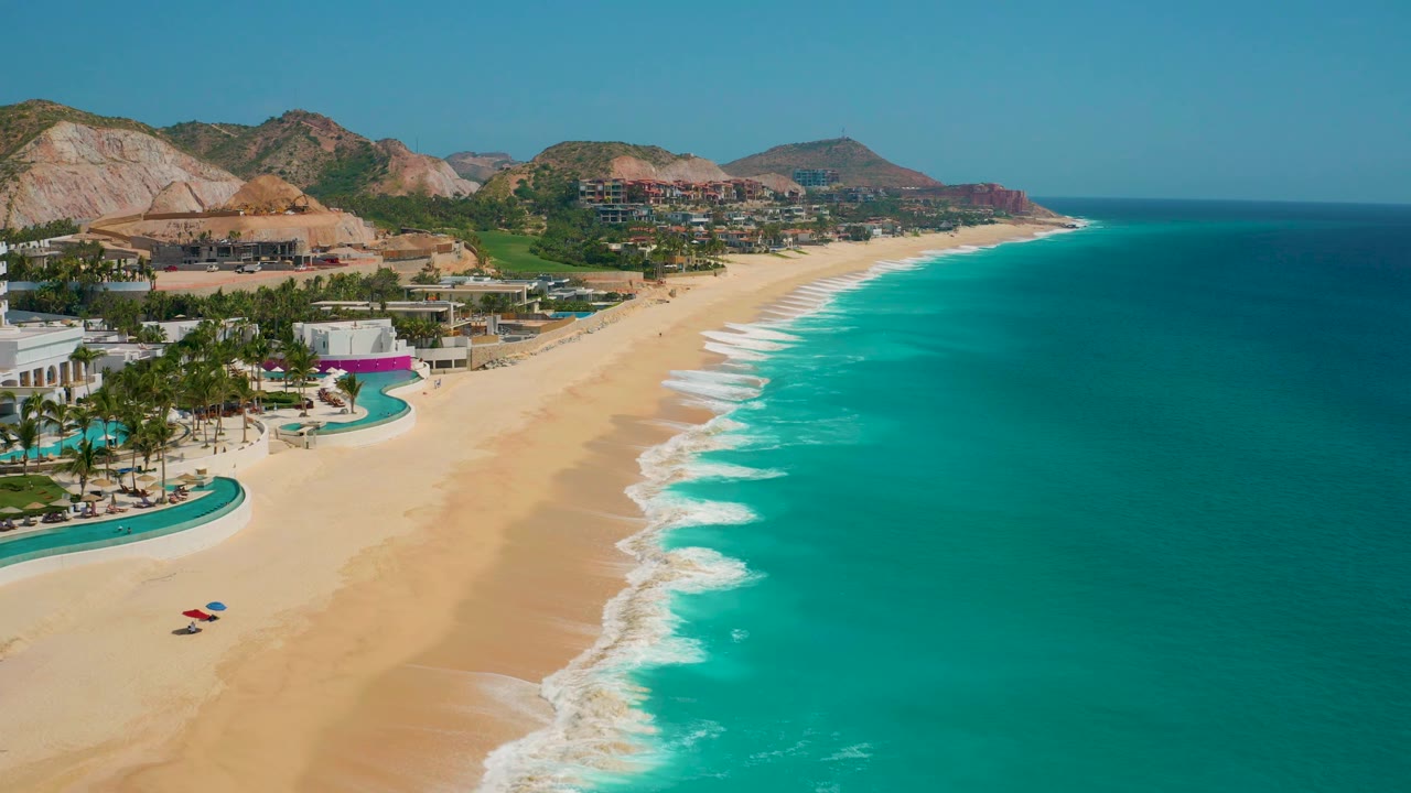 Aerial coastline at Secrets Marquis Los Cabos, Mexico, with two small beach umbrellas on the sand, resort pools at the left edge, turquoise water to the right, and no visible people.