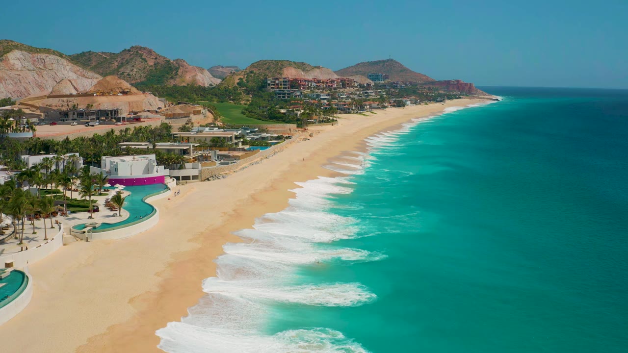 At 0:01 the elevated view keeps the pool and palms in the foreground while white surf bands roll up the Secrets Marquis Los Cabos beach with no visible people.