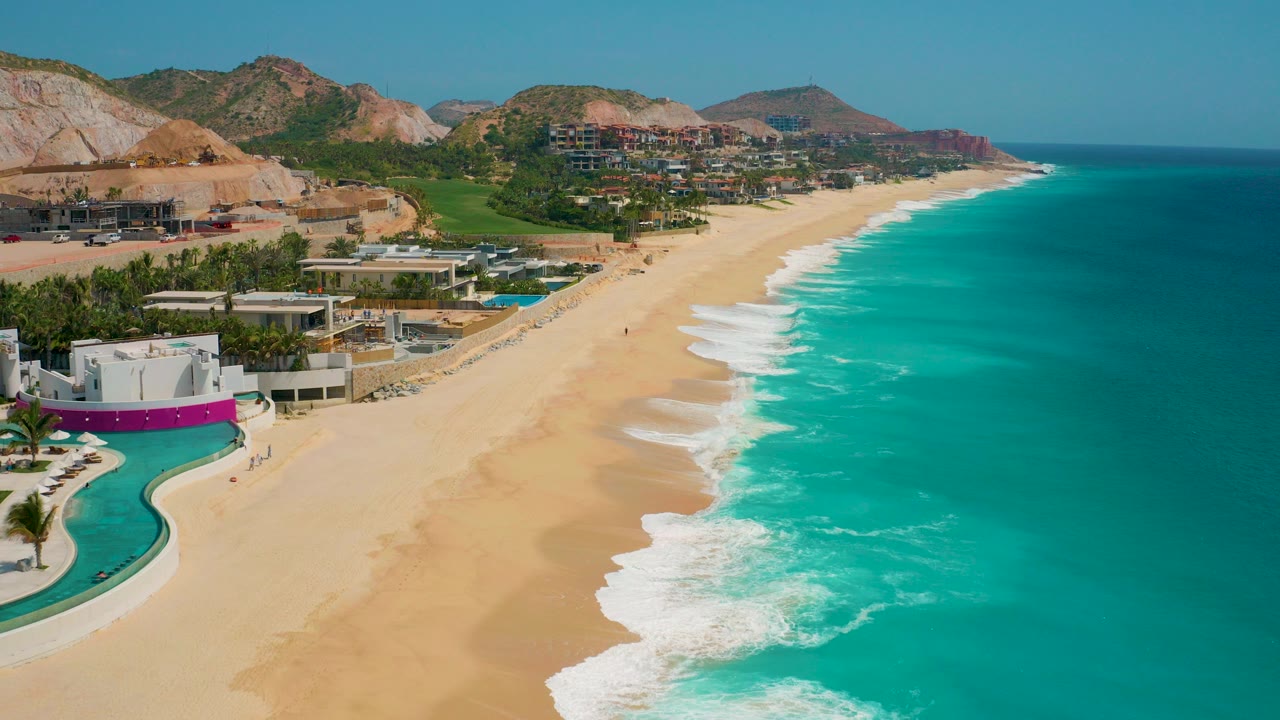 Opening aerial frame over Secrets Marquis Los Cabos in Mexico, with resort pools at the lower left, a broad golden beach, turquoise surf on the right, low hills behind, and no visible people.
