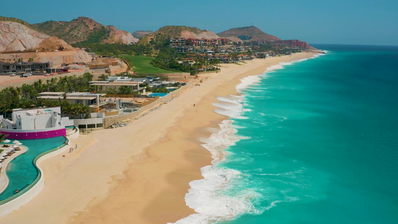 Aerial frame over the Secrets Marquis Los Cabos beachfront in Mexico, with resort pools and walls at left, diagonal white wave lines across the sand, deep blue water offshore, and no visible people.