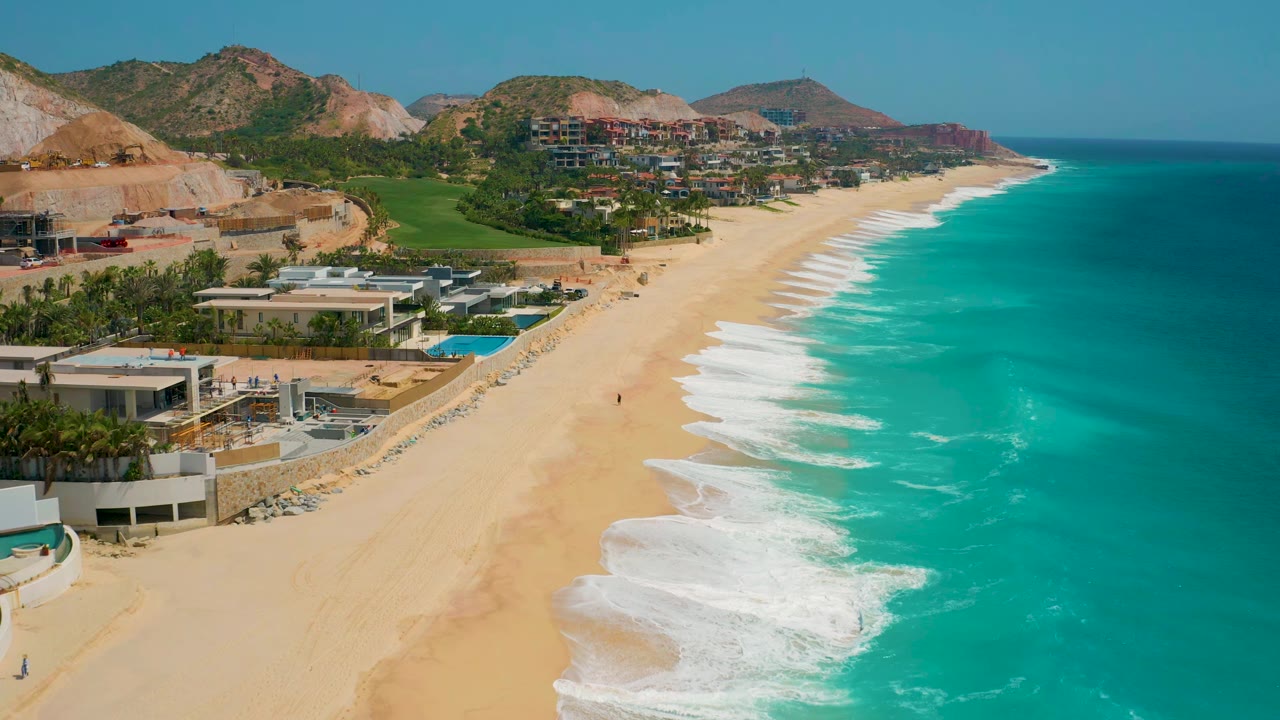 Wide beach and ocean aerial with homes and hills along the shore.