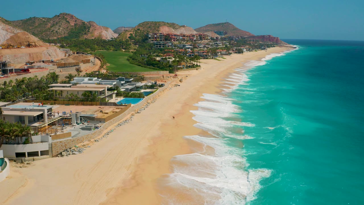 At 0:02, the aerial view holds on the Secrets Marquis Los Cabos coastline with more open ocean to the right, bright surf along the beach, developed lots on the left, and no visible people.