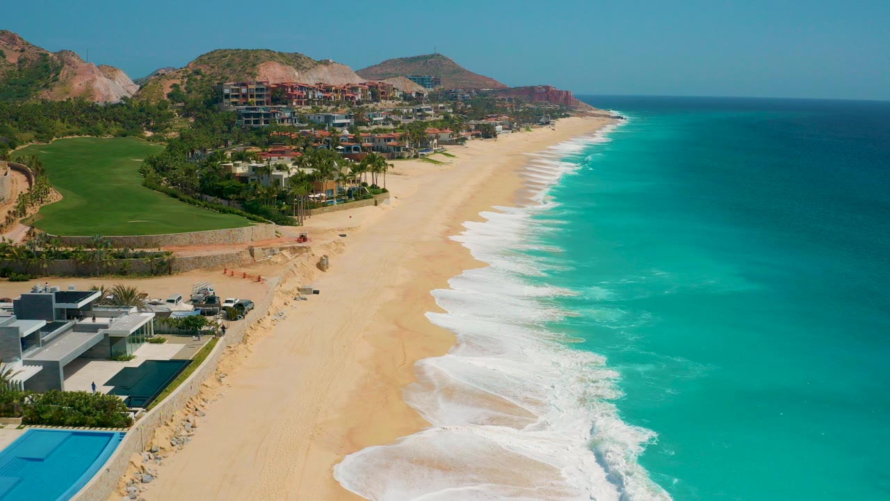 Opening aerial frame of Secrets Marquis Los Cabos in Mexico with a wide sandy beach, turquoise water, resort buildings, green grounds, rocky hills, and no visible people.