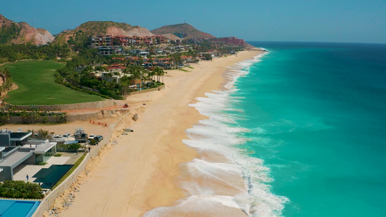 Aerial frame at 0:02 with resort roofs at the lower left, broad sand through the center, blue-green ocean to the right, distant hills, and no visible people.