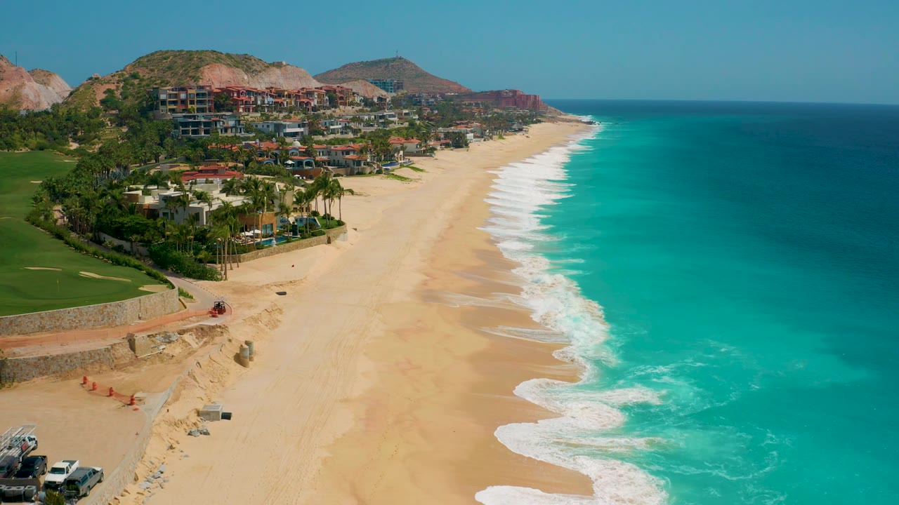 At 0:02, the aerial view favors more open water on the right while the Secrets Marquis Los Cabos beach, hillside buildings, and no visible people remain clear.