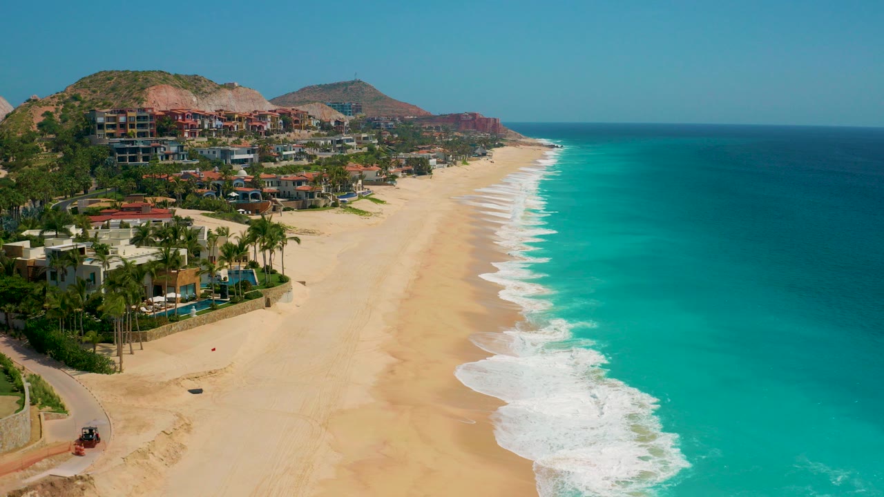 Opening aerial view at Secrets Marquis Los Cabos, Mexico, with resort buildings and palms at left, an empty sandy beach below, turquoise ocean at right, and no visible people.