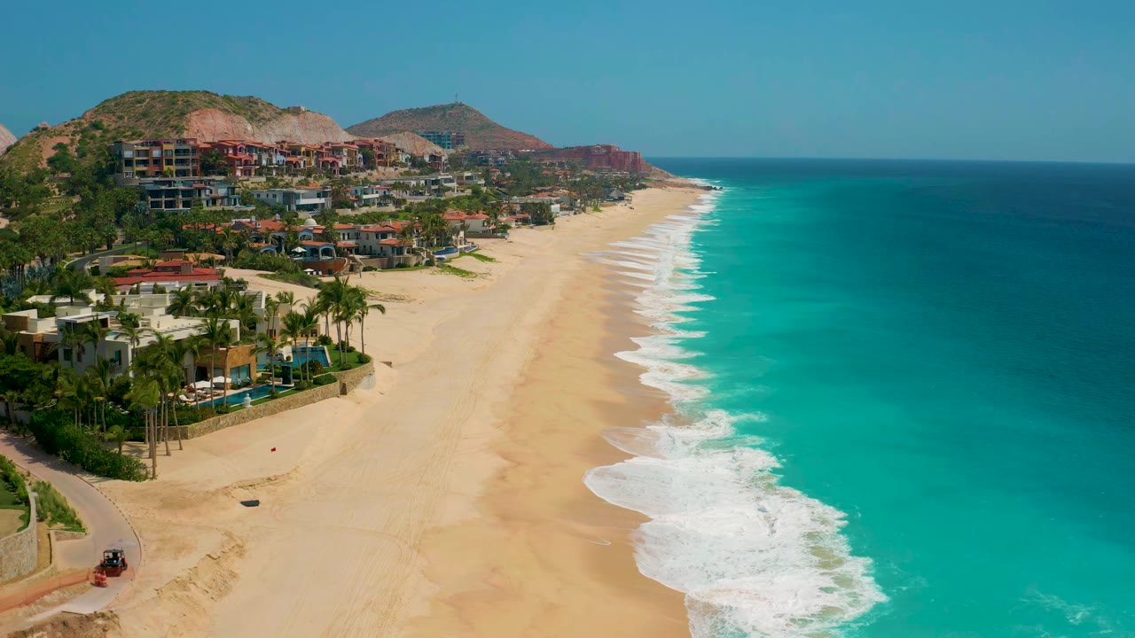 Aerial coastline view at Secrets Marquis Los Cabos, Mexico, showing foamy waves curling along the empty beach, hillside buildings to the left, and no visible people.