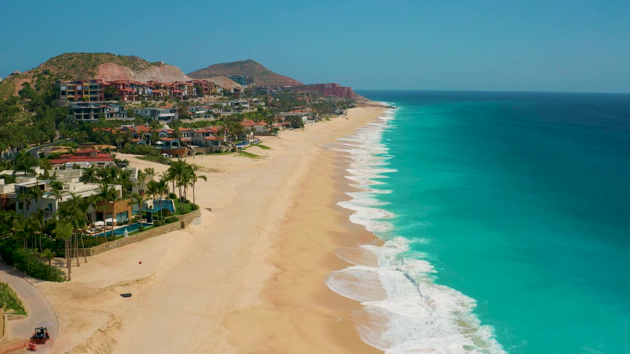 Elevated view of Secrets Marquis Los Cabos, Mexico, with resort grounds and palms in the lower left, a long empty sand beach through the center, blue-green sea to the right, and no visible people.