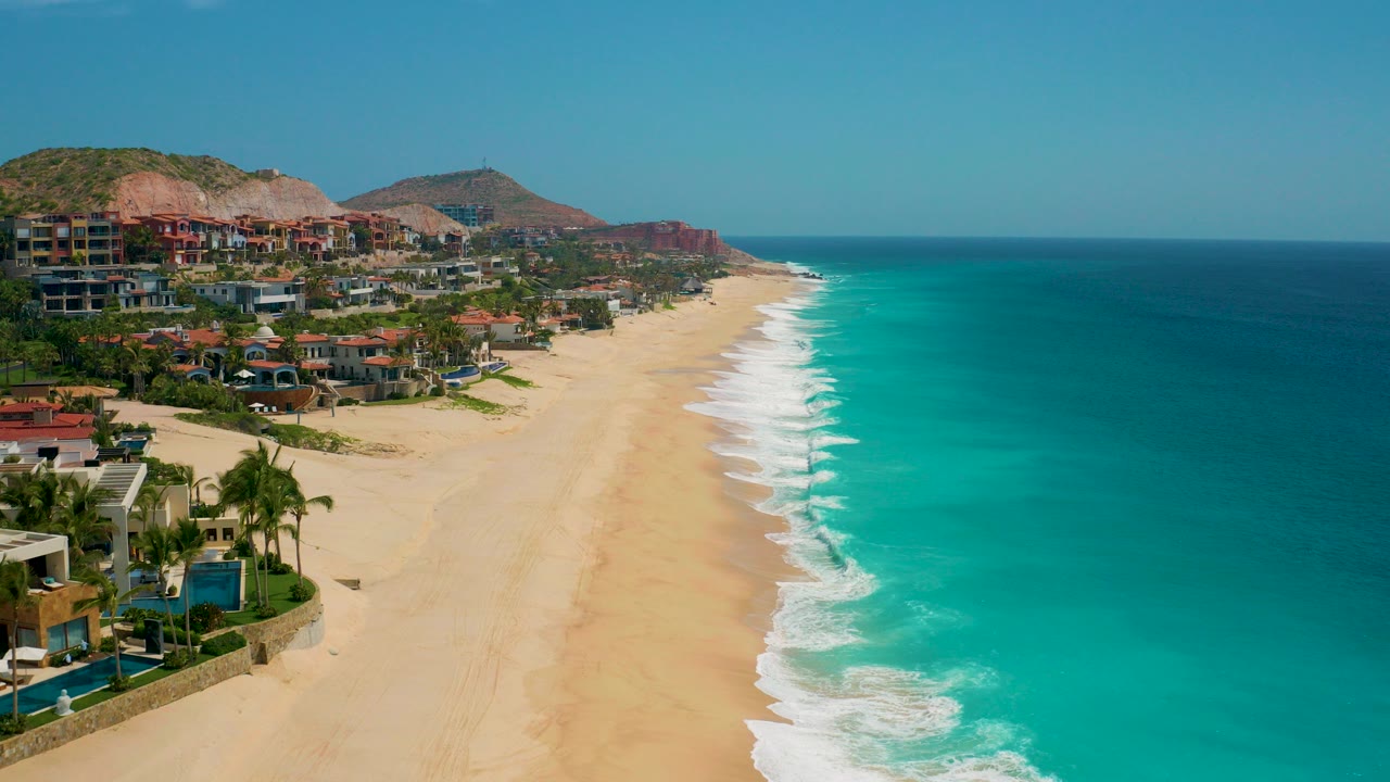 Opening aerial view at Secrets Marquis Los Cabos, Mexico, with the sandy beach running into the distance, turquoise ocean on the right, hillside buildings on the left, and no visible people.