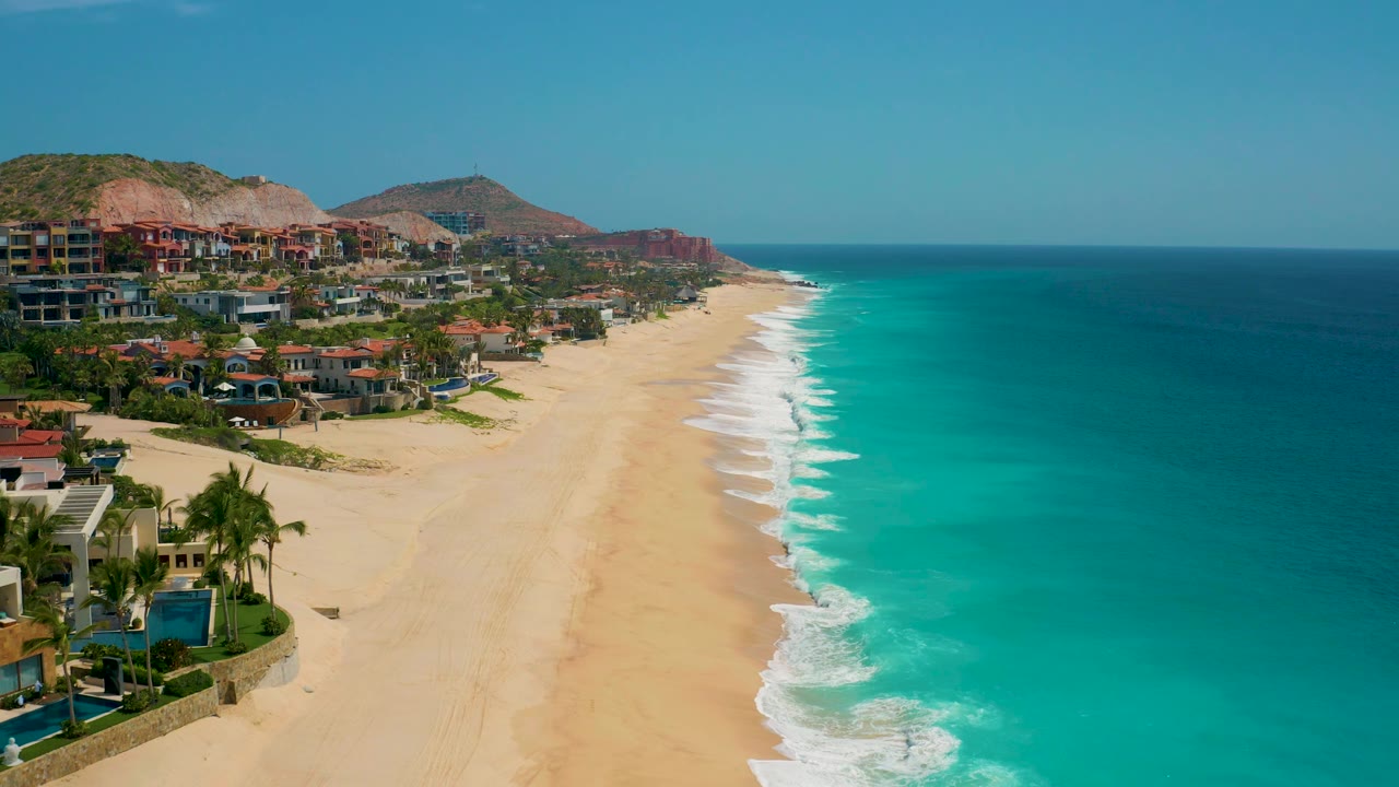 At 0:02, the frame holds a wide Secrets Marquis Los Cabos shoreline with the ocean filling the right side, beach and buildings on the left, reddish hills beyond, and no visible people.