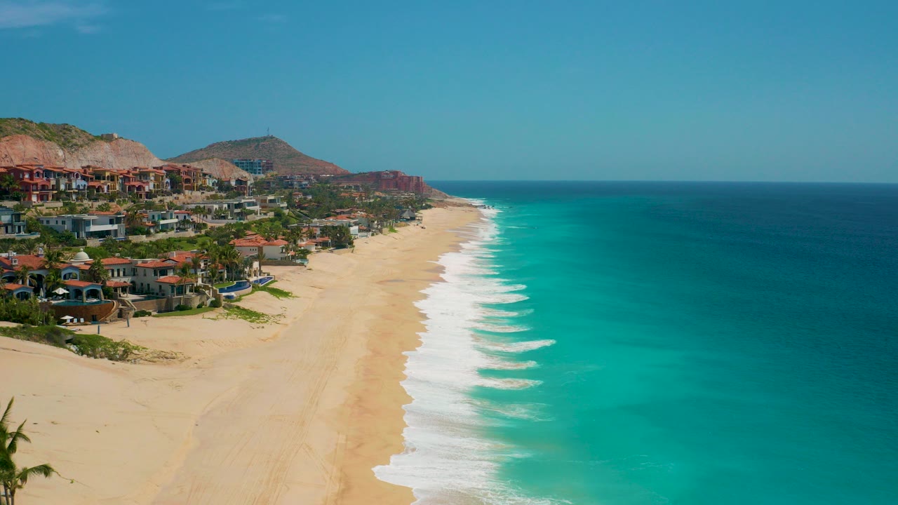 Opening aerial frame over Secrets Marquis Los Cabos, Mexico shows pale sand, turquoise waves, hillside buildings, red-brown mountains, blue sky, and no visible people.