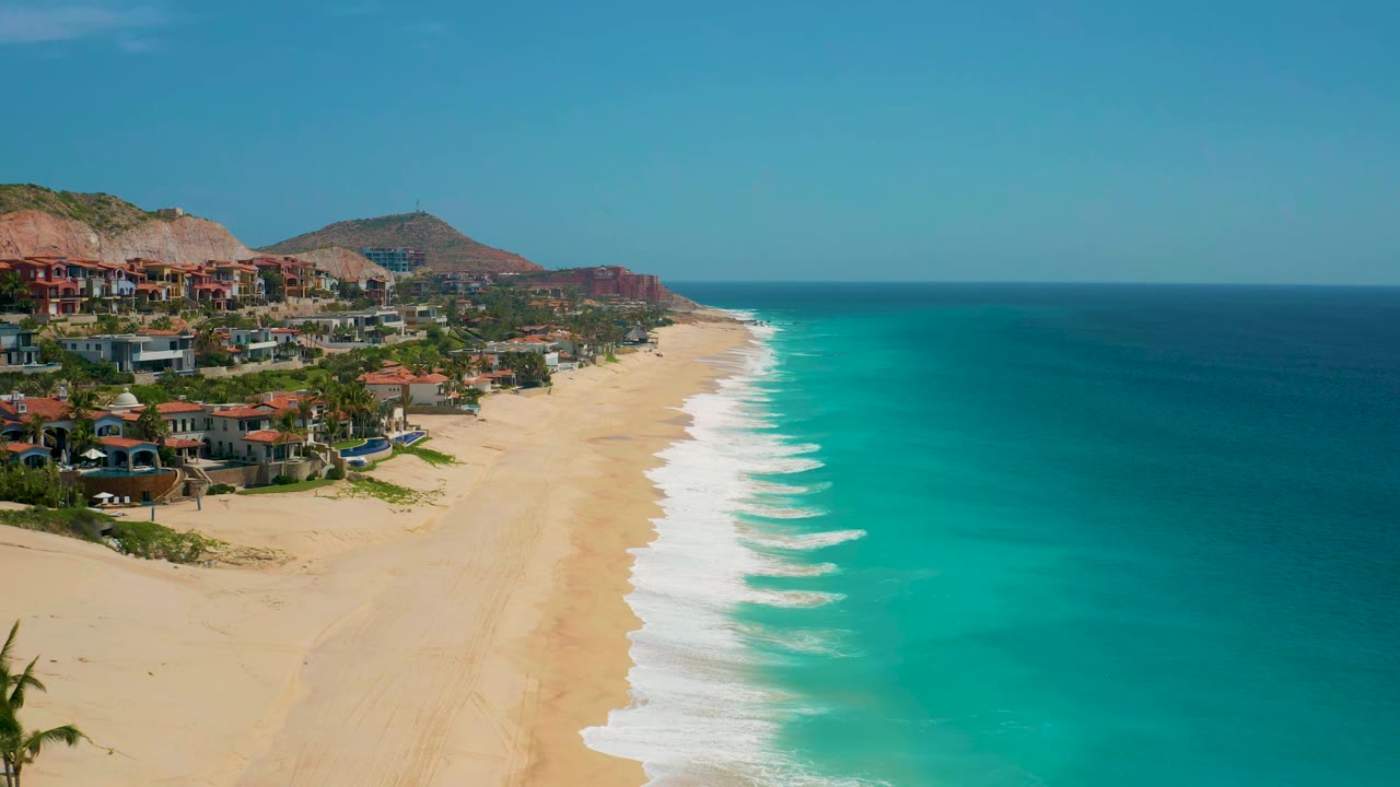At 0:01, the aerial view holds the Secrets Marquis Los Cabos shoreline with white surf bands, open blue water, beach villas or buildings on the slope, and no visible people.