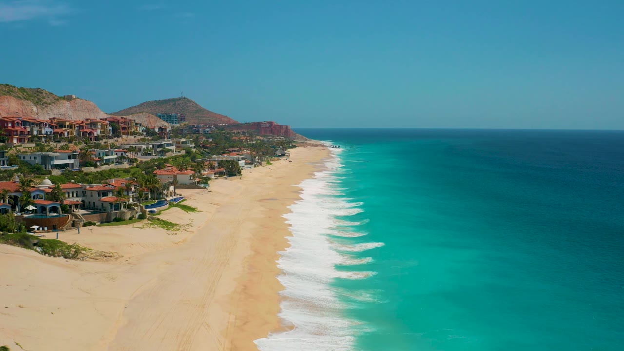At 0:02, the frame keeps the beach and surf centered while deep ocean fills the right side and hillside development remains visible on the left, with no visible people.