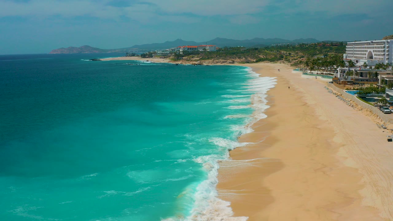 Aerial view at Secrets Marquis Los Cabos, Mexico, with turquoise ocean on the left, white surf reaching broad sand, resort buildings at right, distant hills on the horizon, and no visible people.