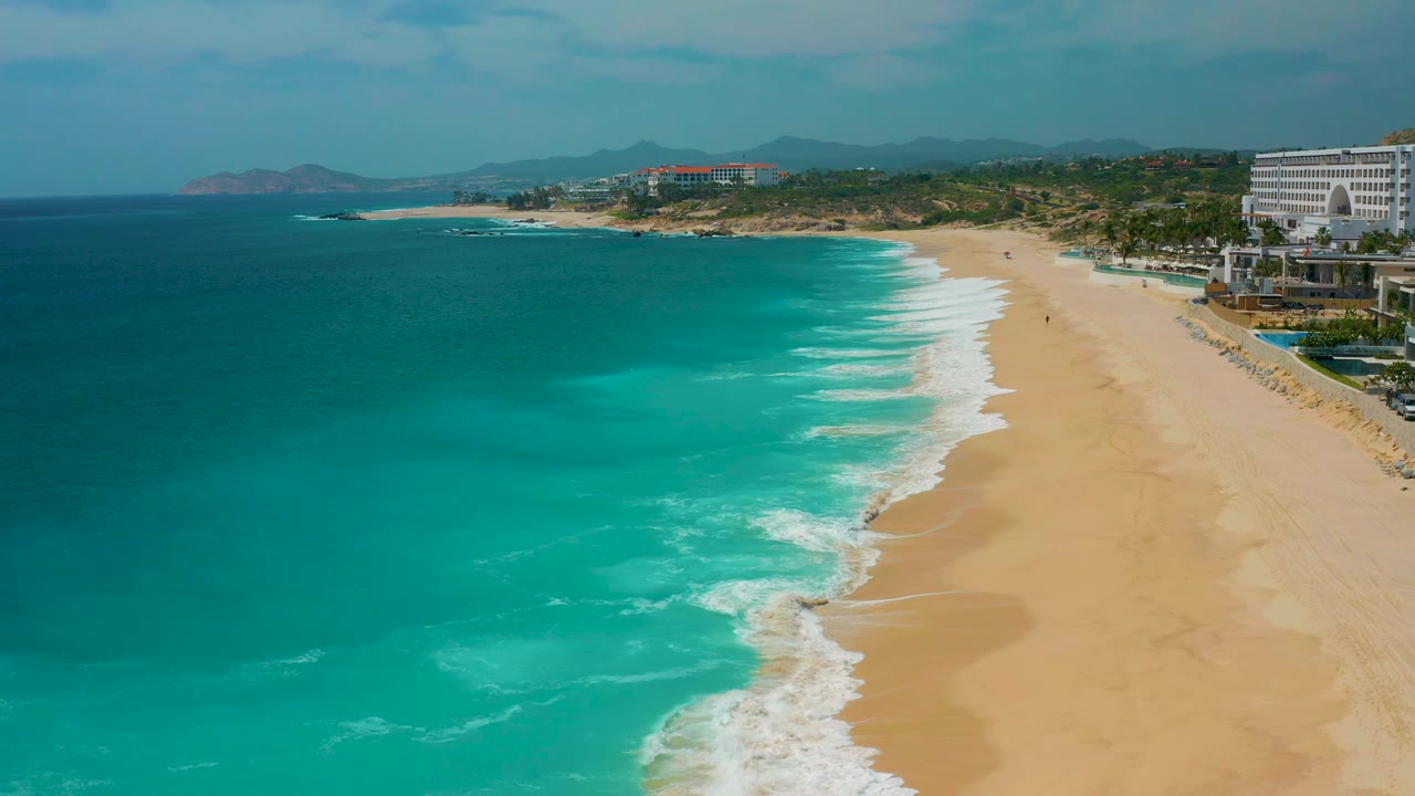 Aerial coastline view at Secrets Marquis Los Cabos, Mexico, with the surf band shifted along the beach, smooth sand occupying the right side, resort buildings near the upper right, and no visible people.