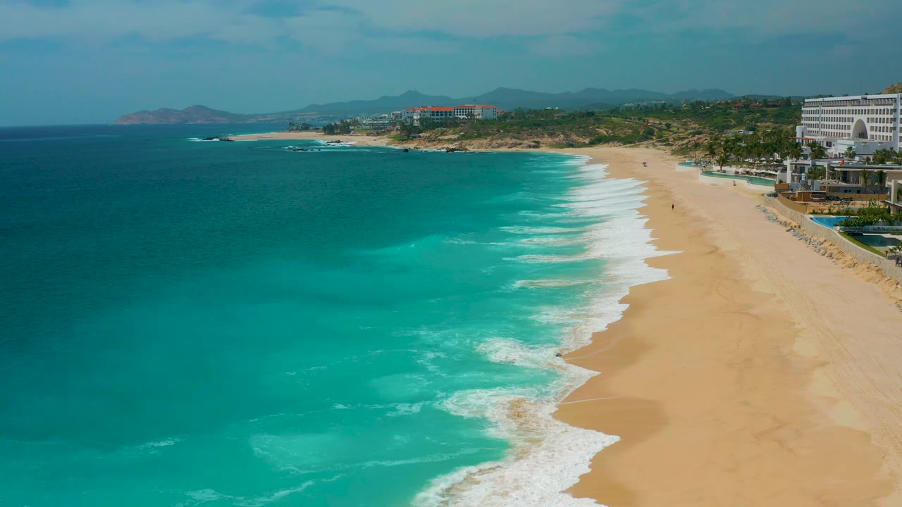 High view at Secrets Marquis Los Cabos, Mexico, with blue-green water taking more of the left frame, a narrow scalloped surf edge on the sand, resort buildings at right, and no visible people.