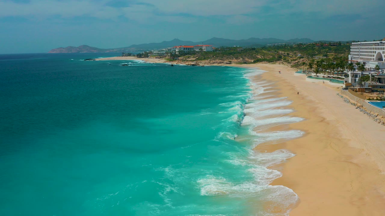 High aerial view over Secrets Marquis Los Cabos, Mexico, with turquoise ocean on the left, pale sand on the right, resort buildings along the shore, distant hills, and no visible people.