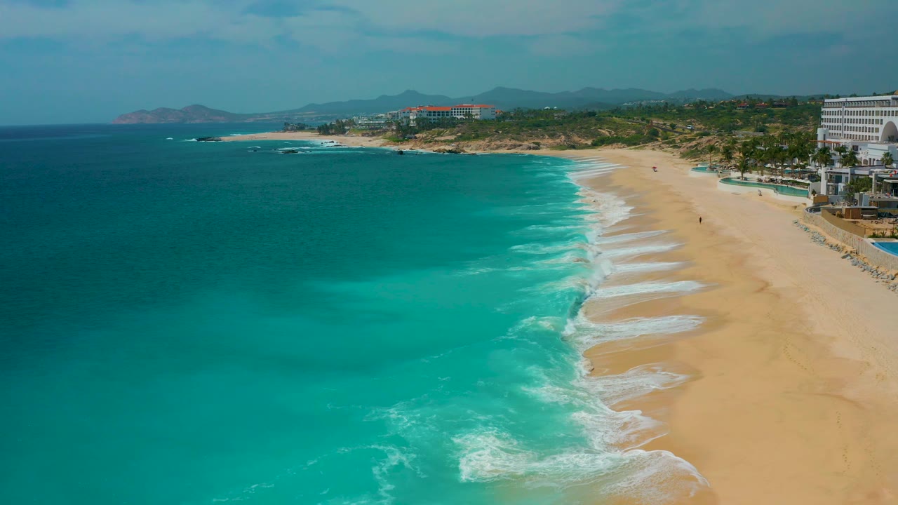 Wide aerial view at Secrets Marquis Los Cabos, Mexico, with more open water filling the left side, white surf curling onto pale sand, resort structures at the right edge, and no visible people.