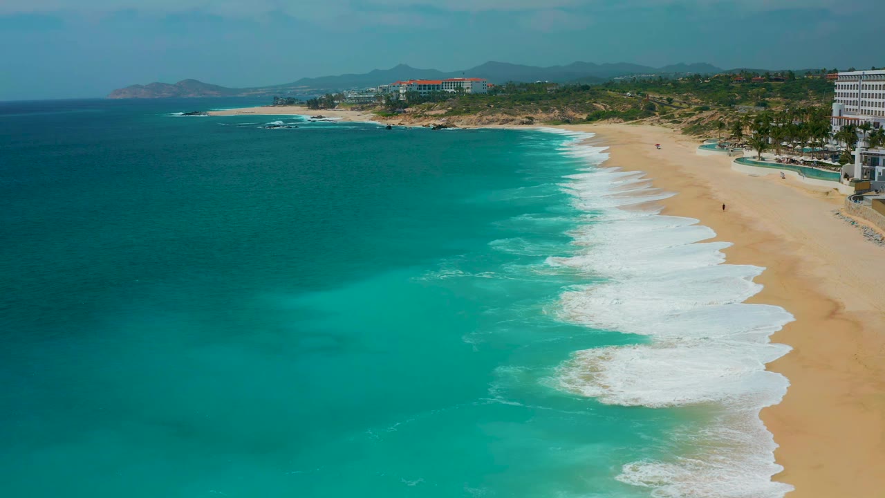 Aerial beach frame at Secrets Marquis Los Cabos, Mexico, with surf lines spread across the sand, deep blue water offshore, and no visible people.