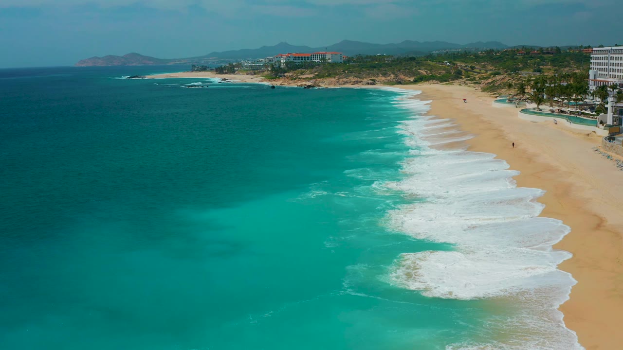 Aerial view of Secrets Marquis Los Cabos, Mexico, with bright turquoise shallows, foamy shore break, wide sand, hillside background, and no visible people.