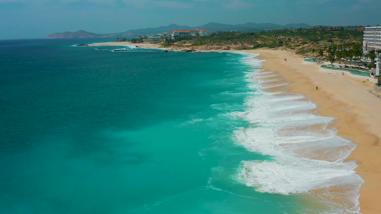 Aerial coastline at Secrets Marquis Los Cabos, Mexico, showing darker offshore water, lighter shallows, white surf, wide sand, and no visible people.