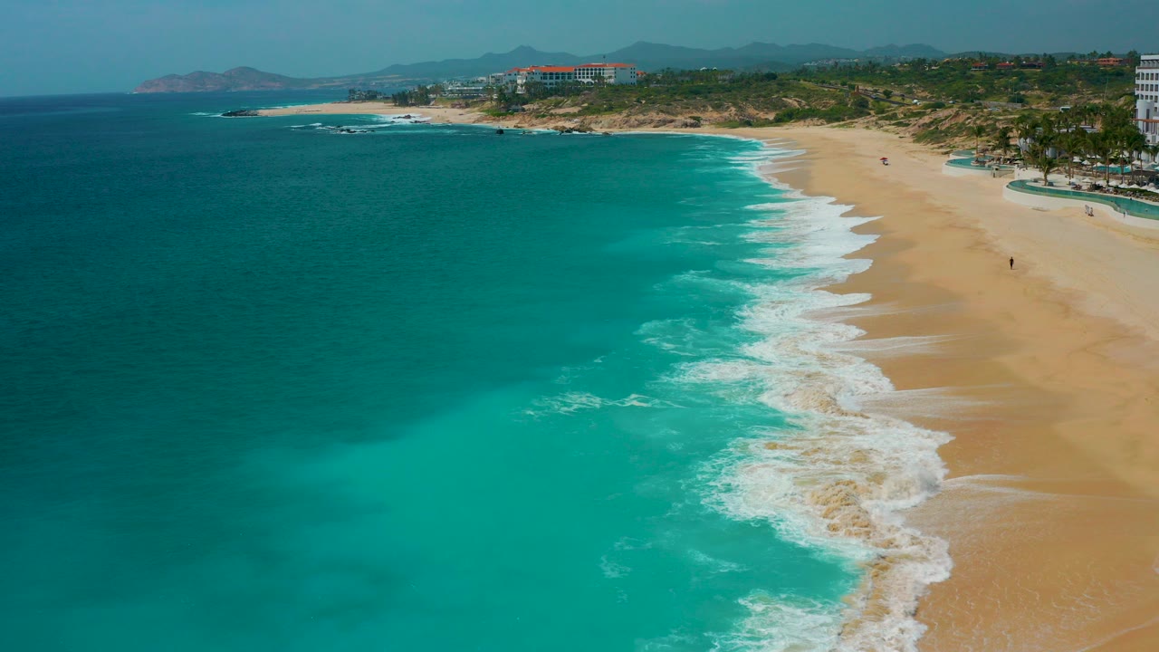Aerial view over Secrets Marquis Los Cabos, Mexico, with turquoise ocean on the left, white surf curling onto a sandy beach on the right, and no visible people.