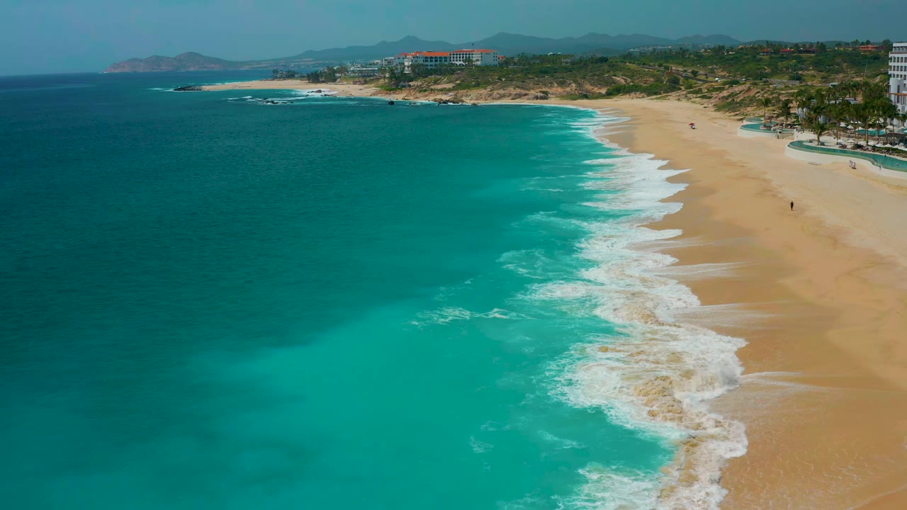Aerial shoreline frame from Secrets Marquis Los Cabos, Mexico, showing white foam advancing farther down the sandy beach with blue-green water filling the left side.
